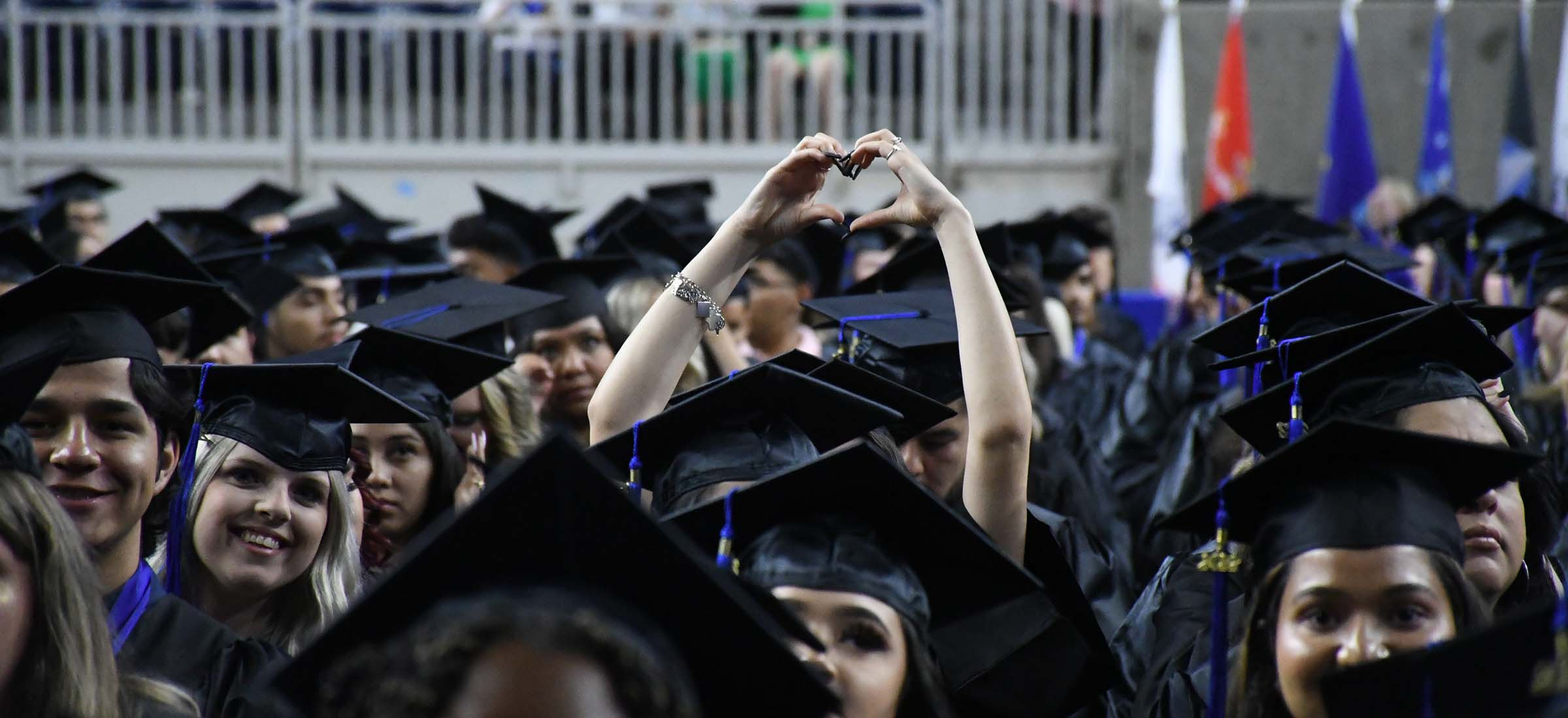 San Jacinto College graduate in cap and gown making a heart gesture with her hands raised in the air during the graduation ceremony